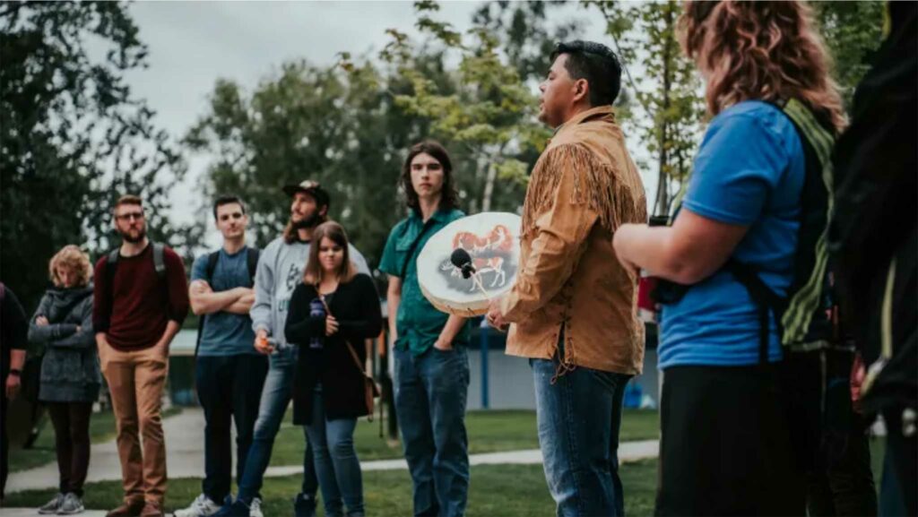 Education students watch as an Indigenous traditional drummer drums in a local park. 